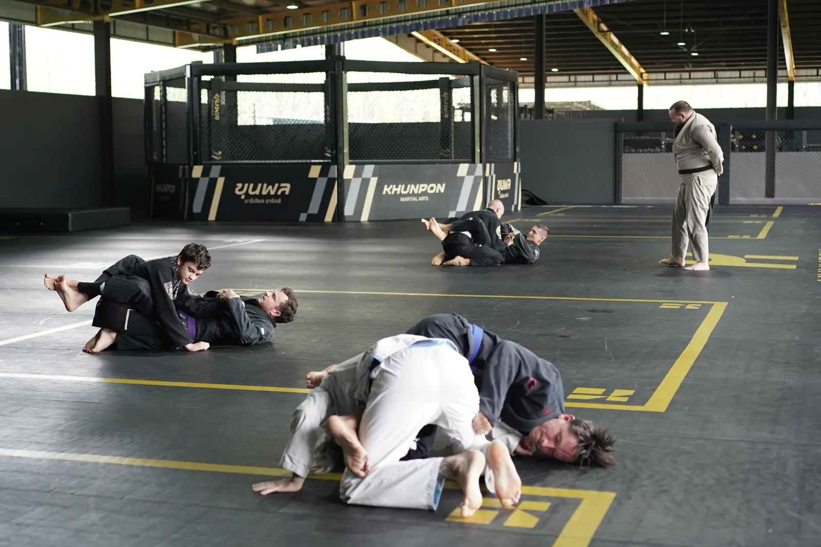 Pairs in black and white gis roll across the Khunpon mats while a coach watches, with the cage at the far end of the hall