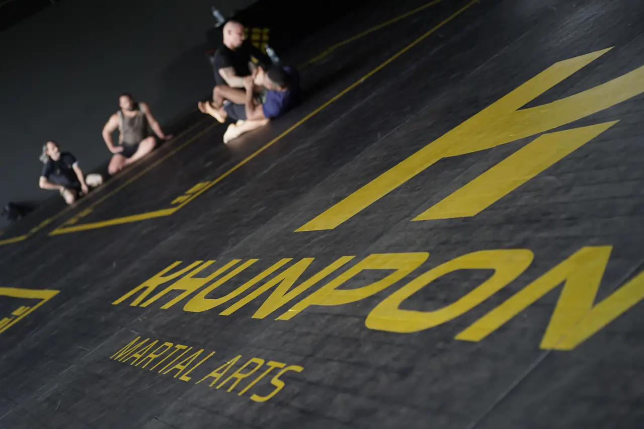 Pairs grapple across the black Khunpon mat, with the gym's logo printed large in gold
