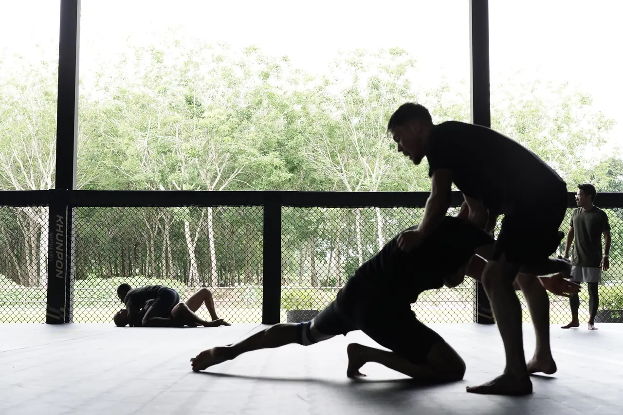 A grappler steps in to control his partner at the open side of the gym, with trees outside