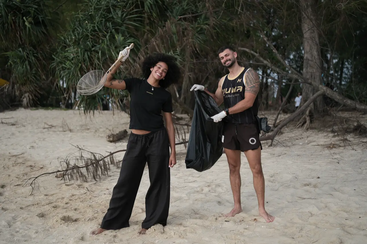 Two Khunpon crew members smiling on the sand, one holding up a full rubbish bag