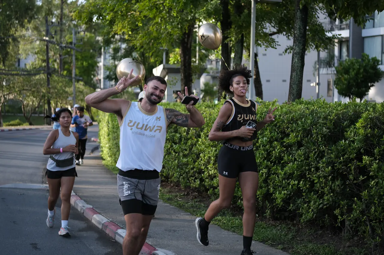 Khunpon crew members running the 5K route beside a hedge, one waving at the camera