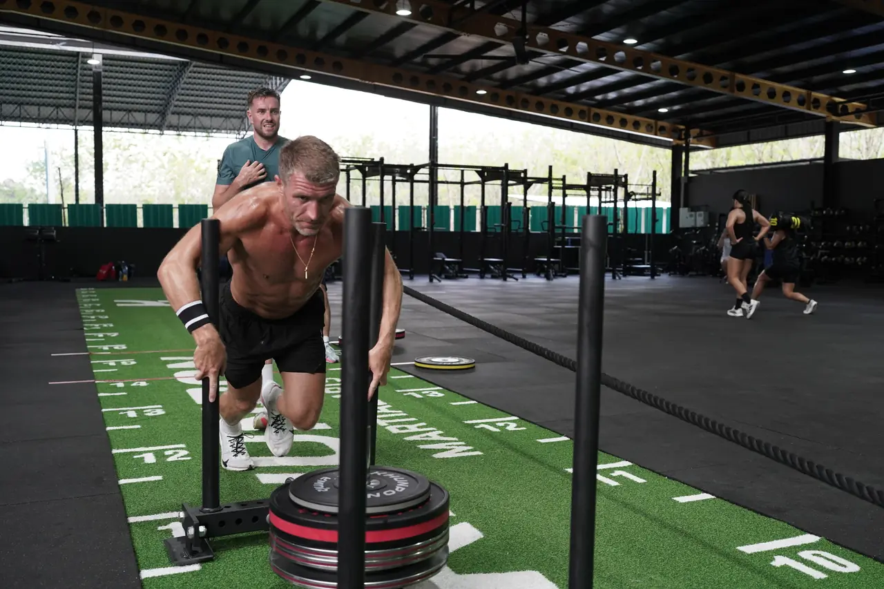 A shirtless athlete leans into the sled push on the turf lane
