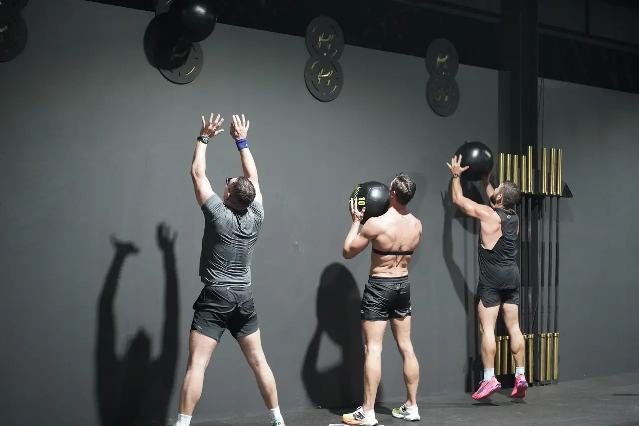 Three athletes throw wall balls against the wall at the final station, their shadows on the concrete