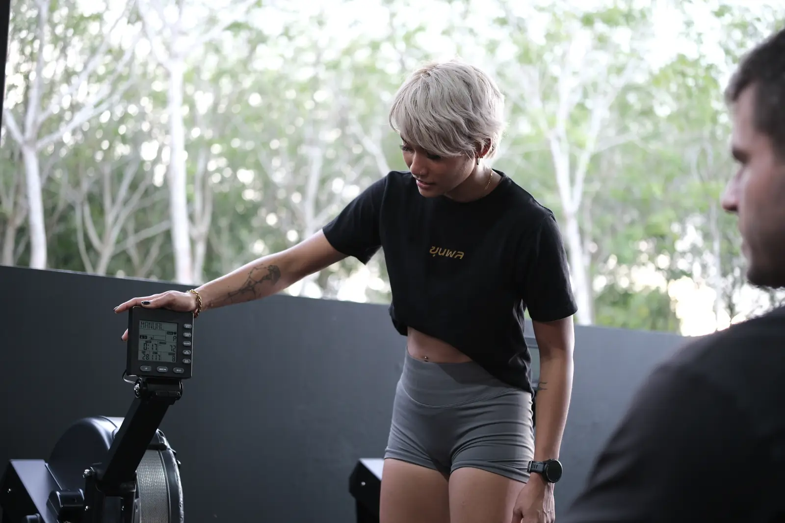 A member checks her rowing machine at the open side of the Khunpon fitness area, with tall trees outside