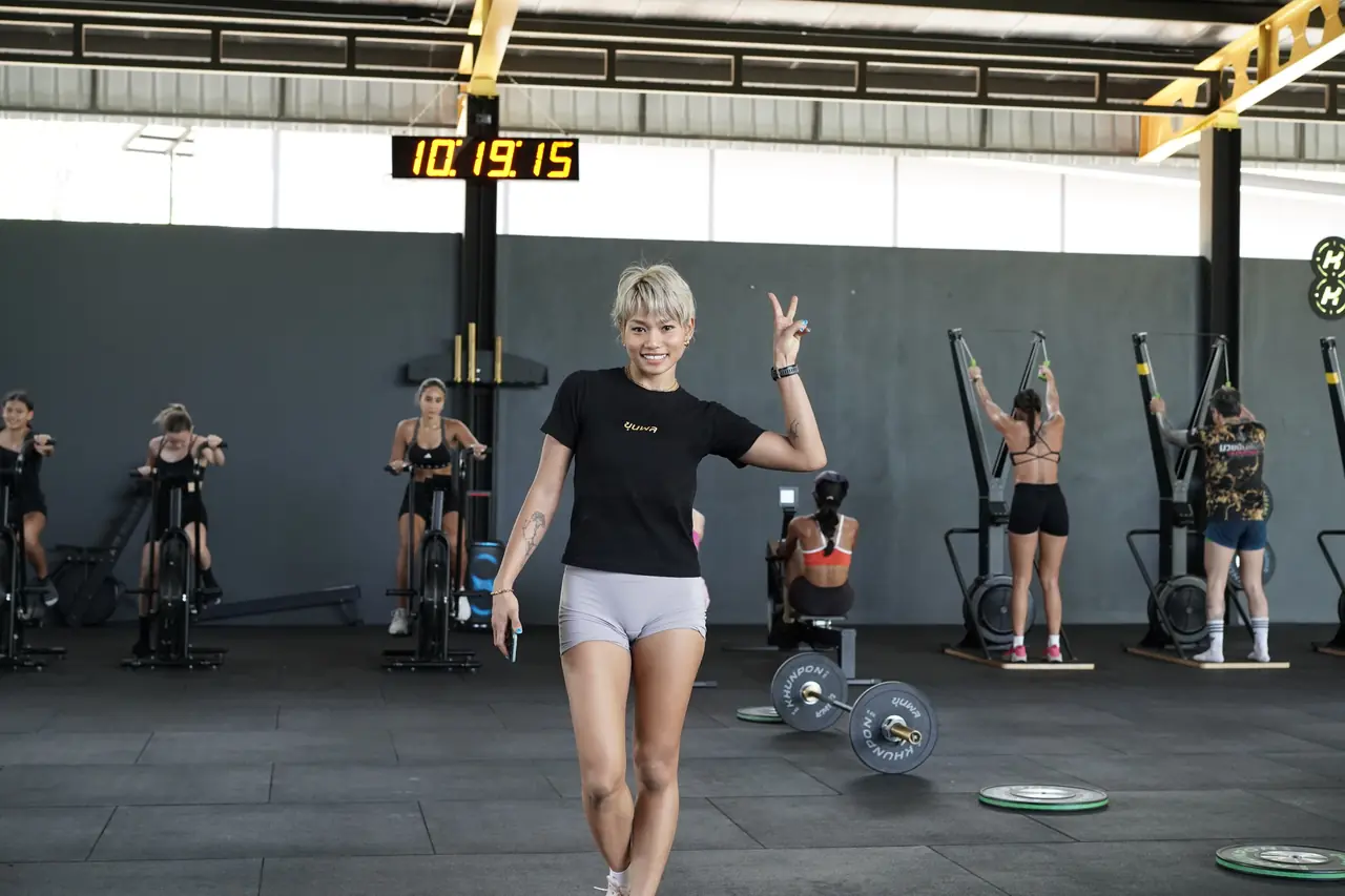 A HYROX coach smiles and waves at the camera as members work on the machines behind her