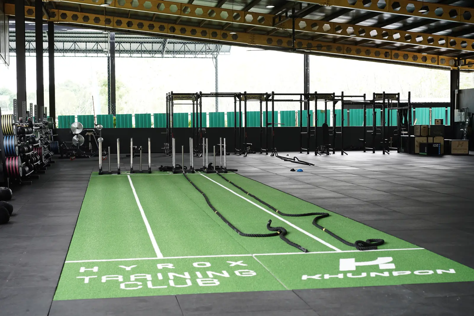 The green HYROX Training Club turf lane at Khunpon, with sleds and battle ropes on it and the racks and machines behind
