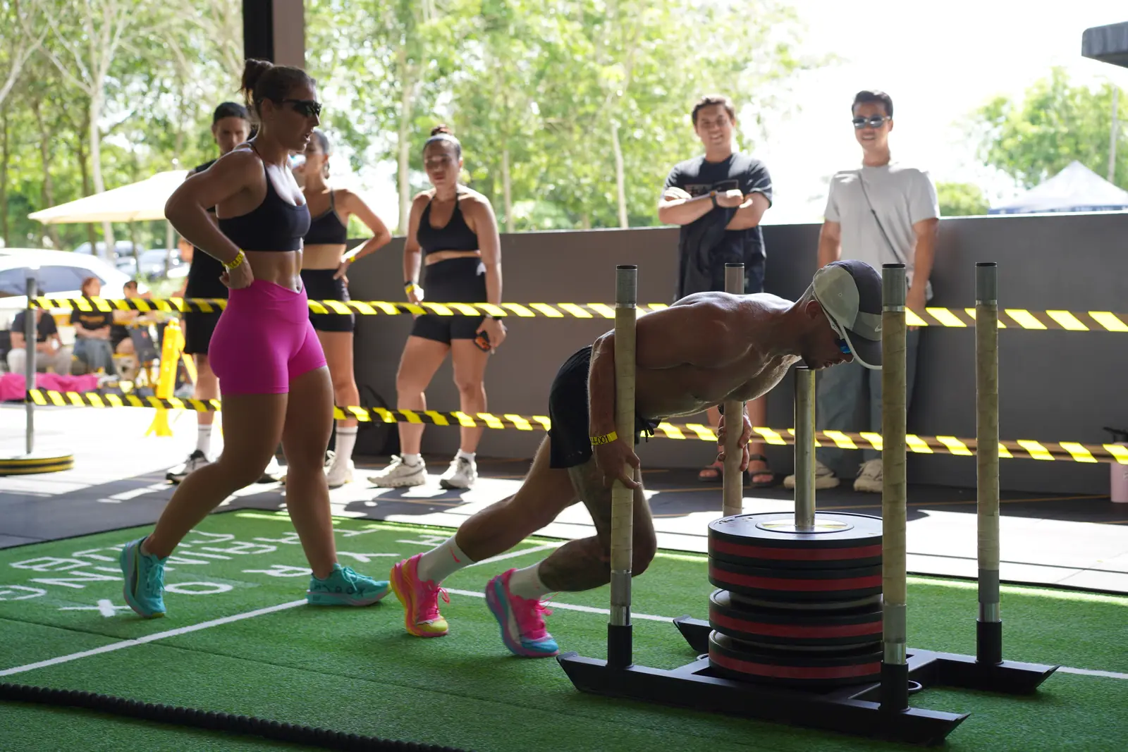 An athlete drives a weighted sled along the turf lane at Khunpon while competitors run past and spectators watch