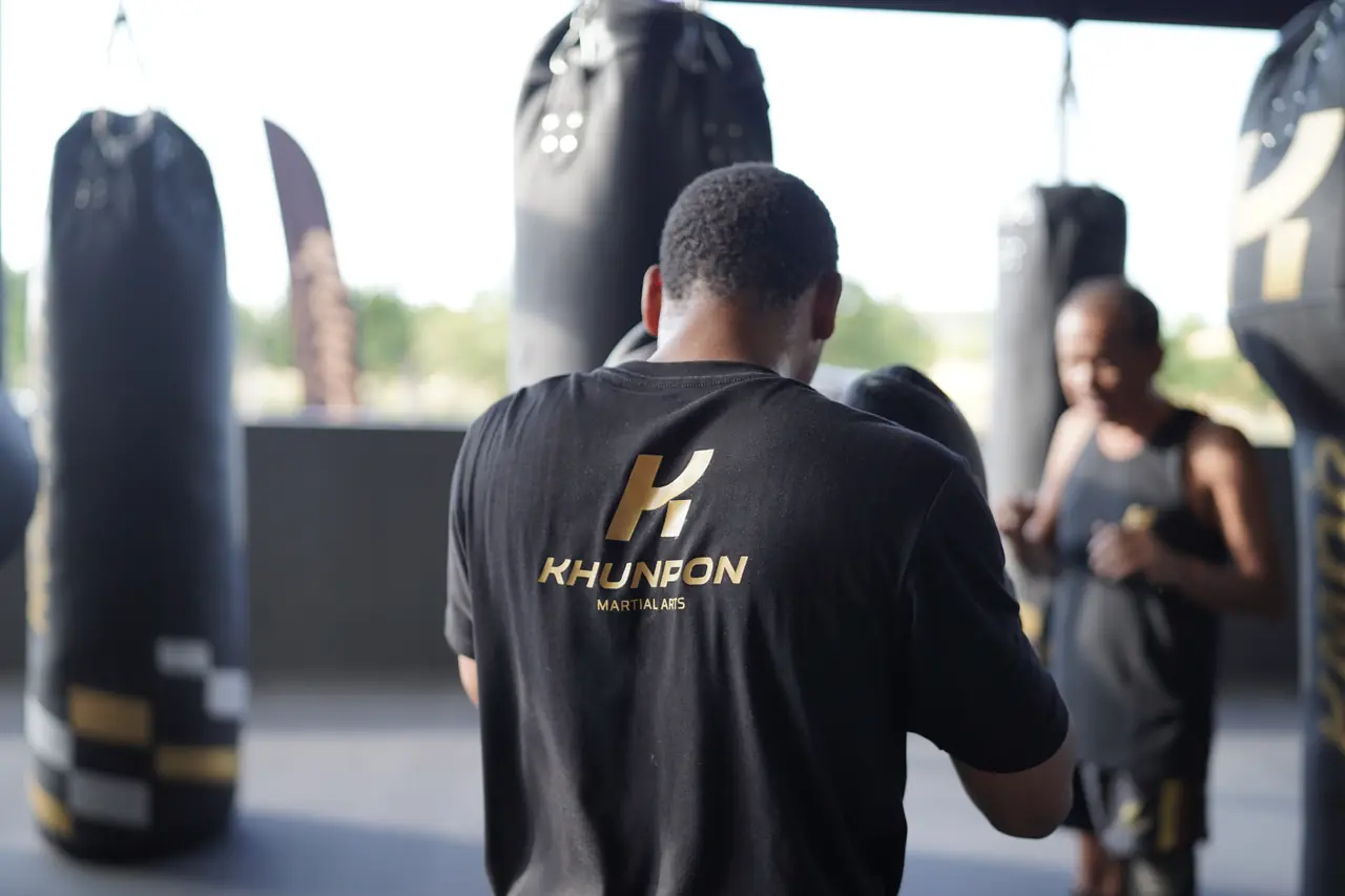 A fighter in a Khunpon shirt faces the heavy bags as the afternoon light comes through the hall