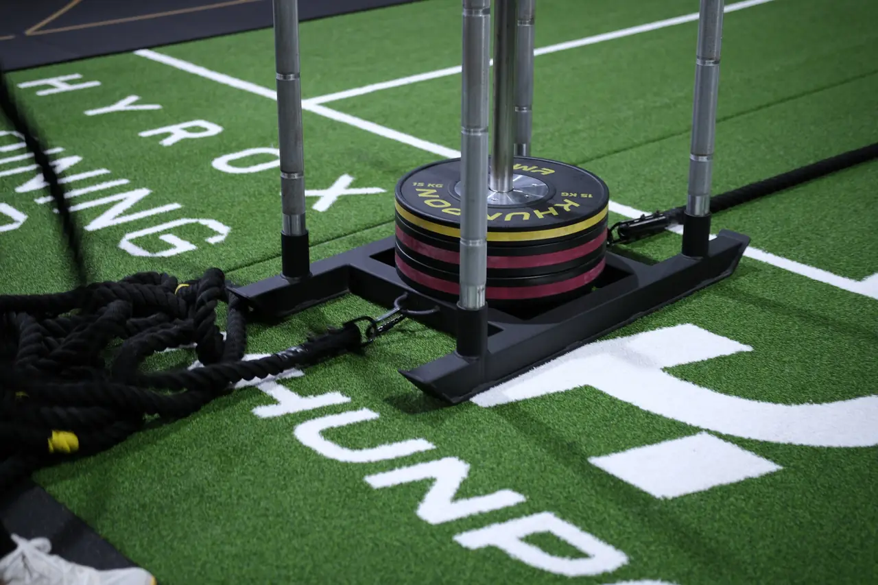 A loaded sled sits on the green turf lane marked HYROX Training Club, with battle ropes beside it