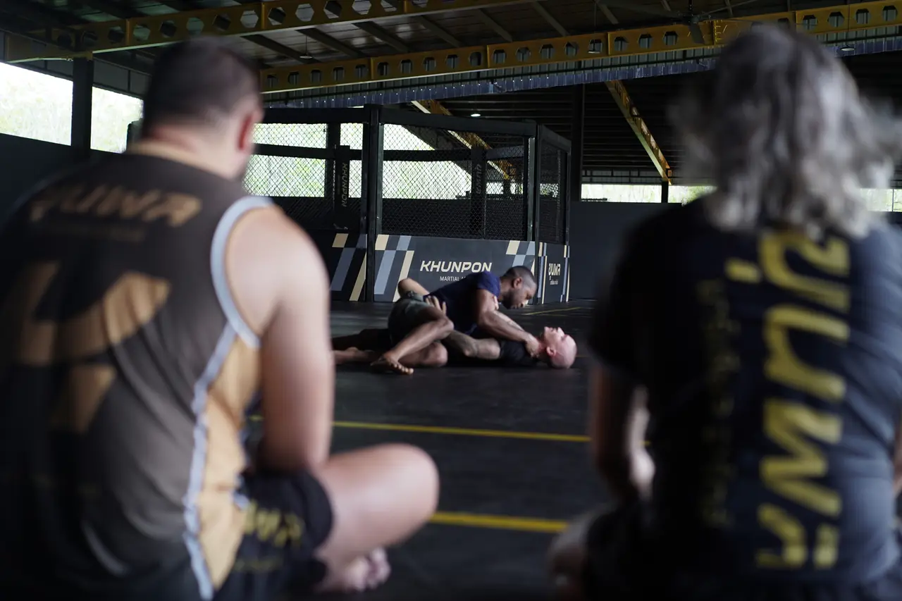 BJJ students sit at the edge of the mats watching a round, with the cage behind them