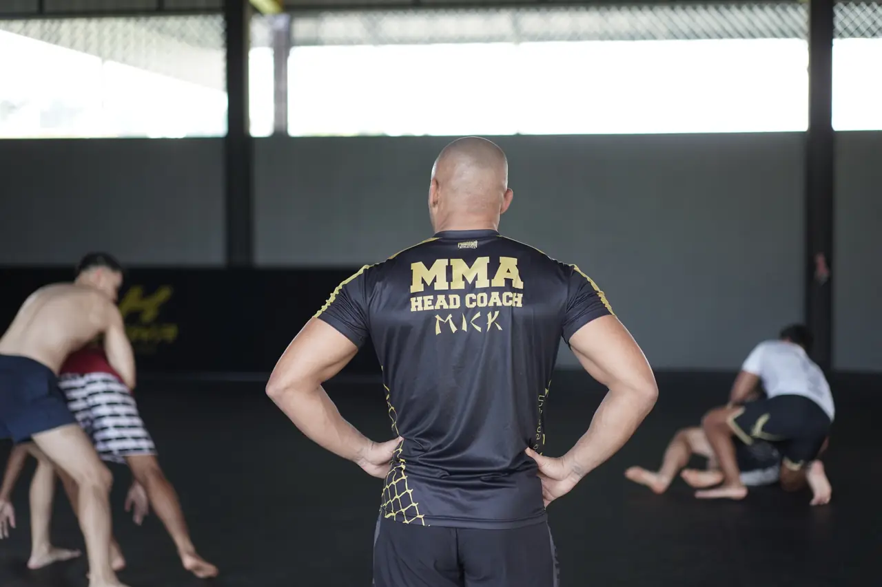 MMA head coach Mick watches from the mats as students grapple in the background