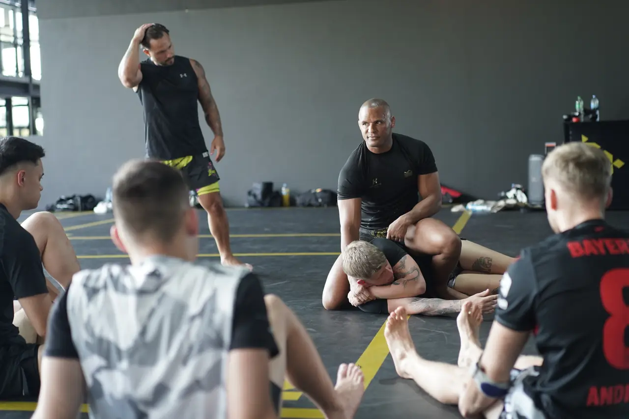 A coach demonstrates a ground position with a partner while students kneel around them on the mats