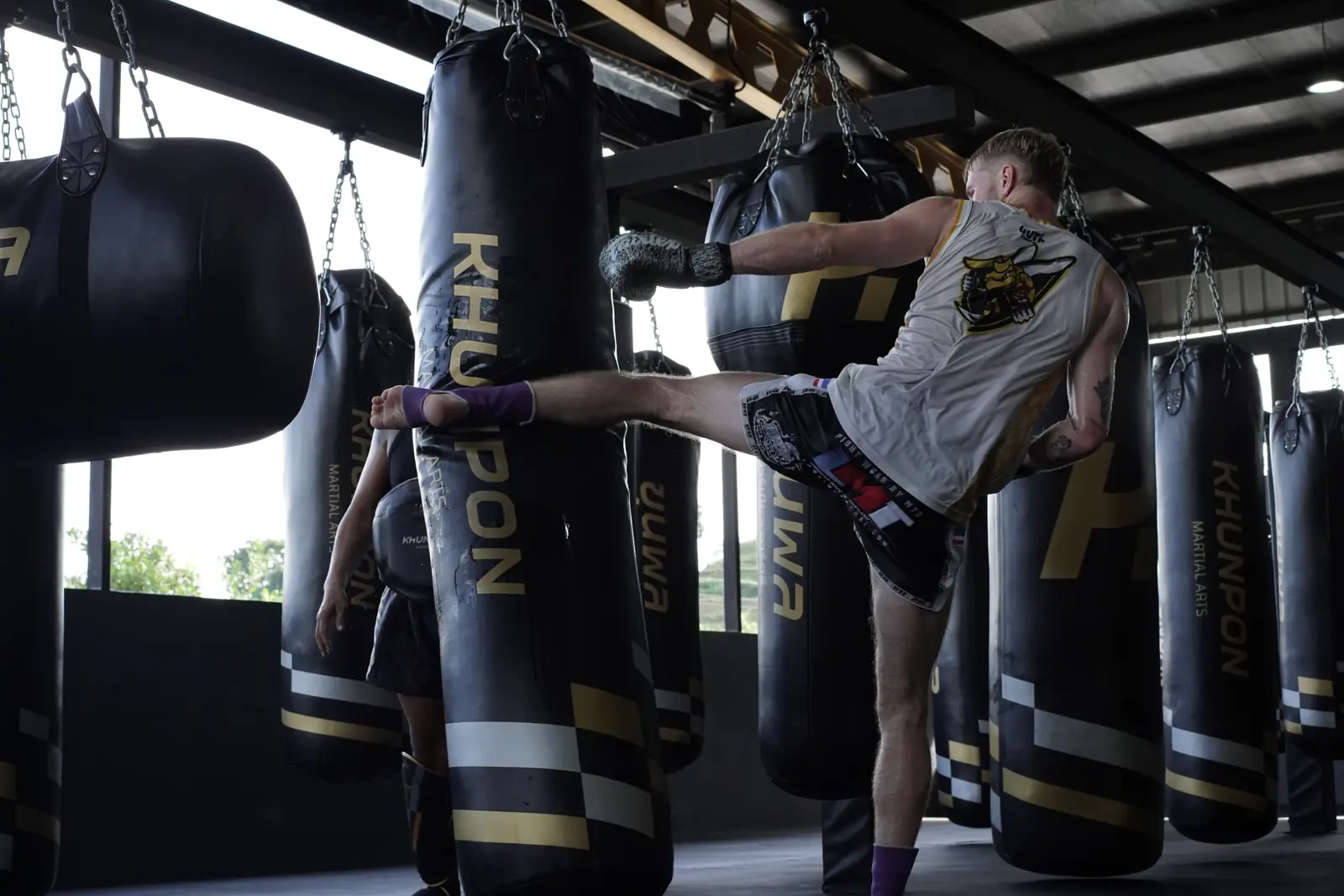 A fighter throws a high roundhouse kick into a heavy bag at Khunpon, with rows of black bags behind him