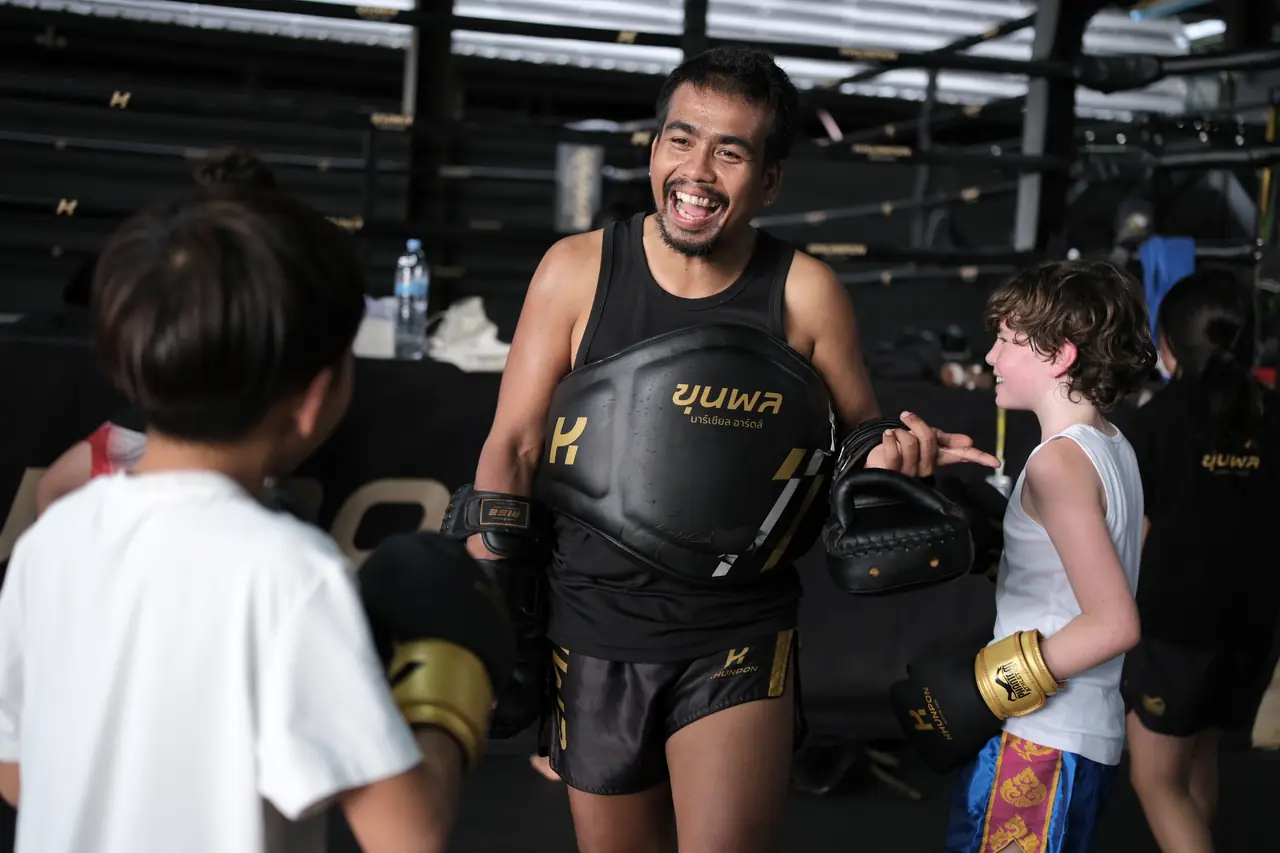 A laughing coach holds a belly pad and focus mitts for young students in a kids' session