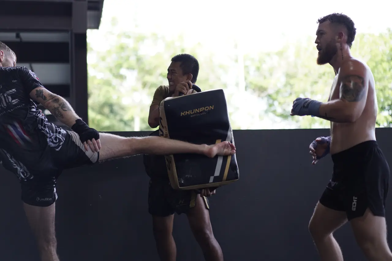 A coach holds a kick shield as a student lands a roundhouse kick on the open-sided training floor