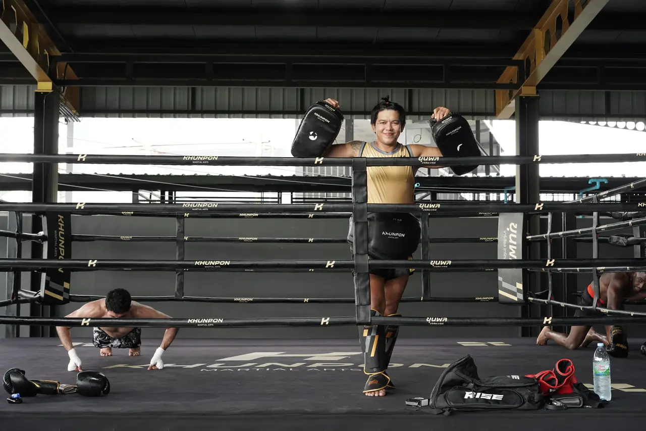 A coach in a gold top stands in the ring holding pads while a fighter does push-ups on the canvas
