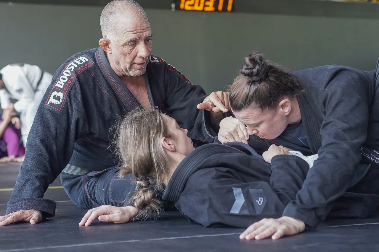 The Professor coaches two women through a position, kneeling beside them on the mat