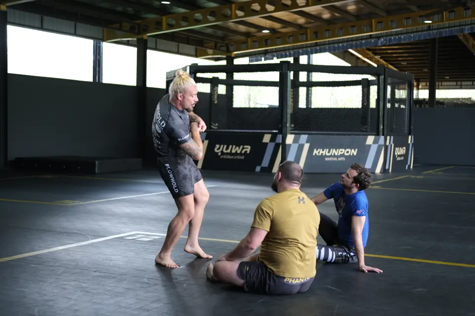 Sloan demonstrates over a partner on the mat while the group watches from the cage side