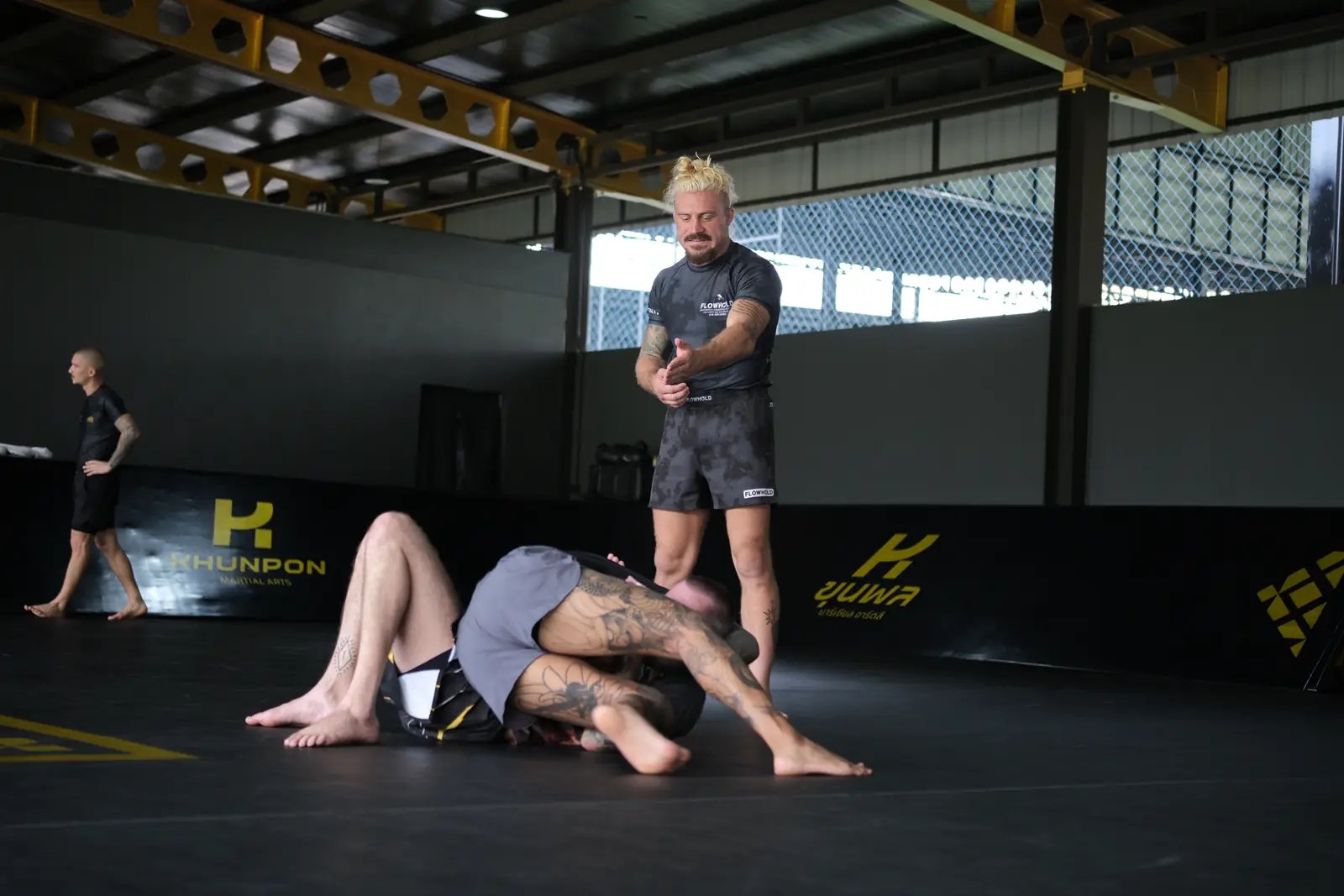 Sloan Clymer stands over a partner on the Khunpon mats during the no-gi seminar, with the open-sided gym behind him