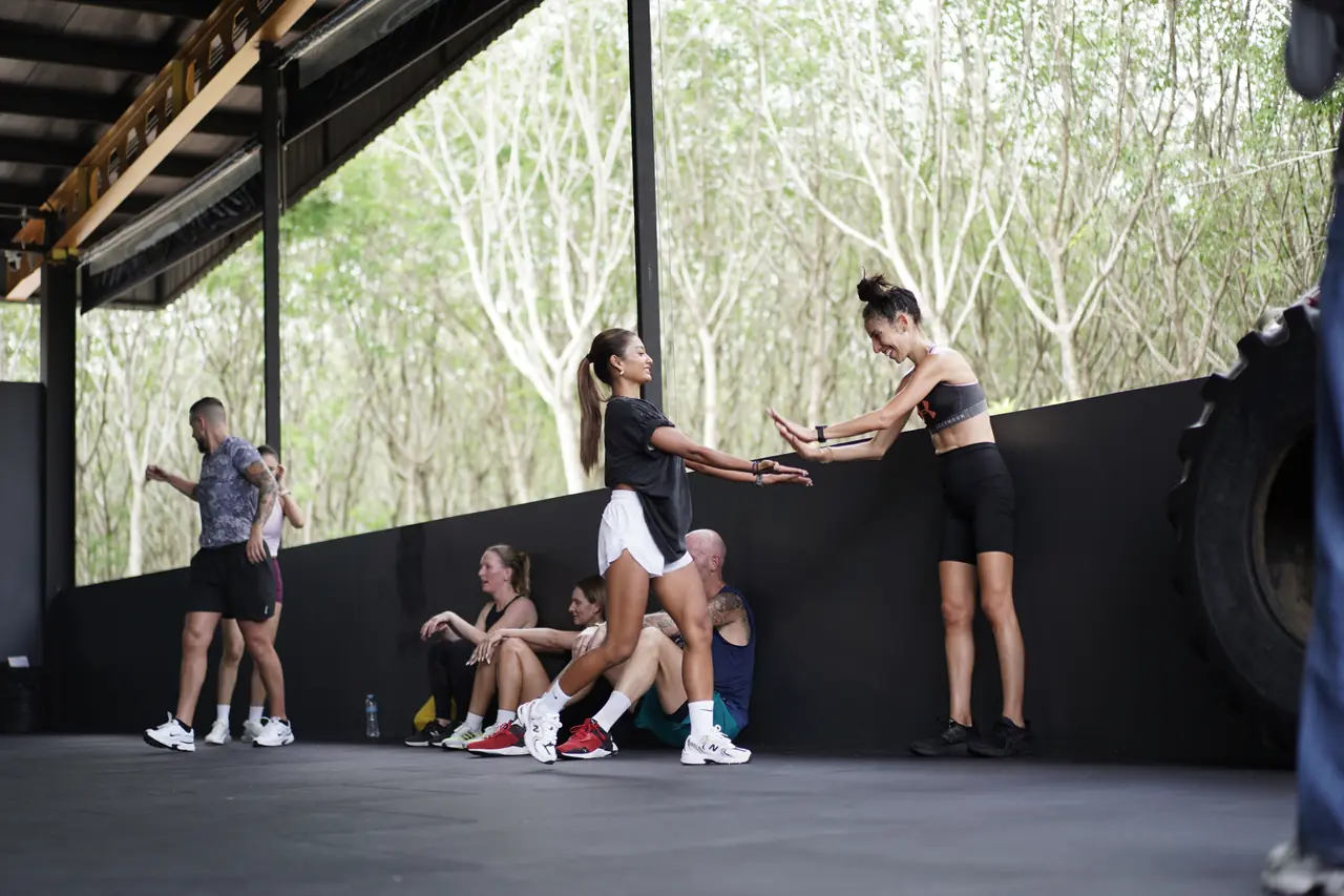 Members stretch together against the wall at the open side of the gym, with trees behind