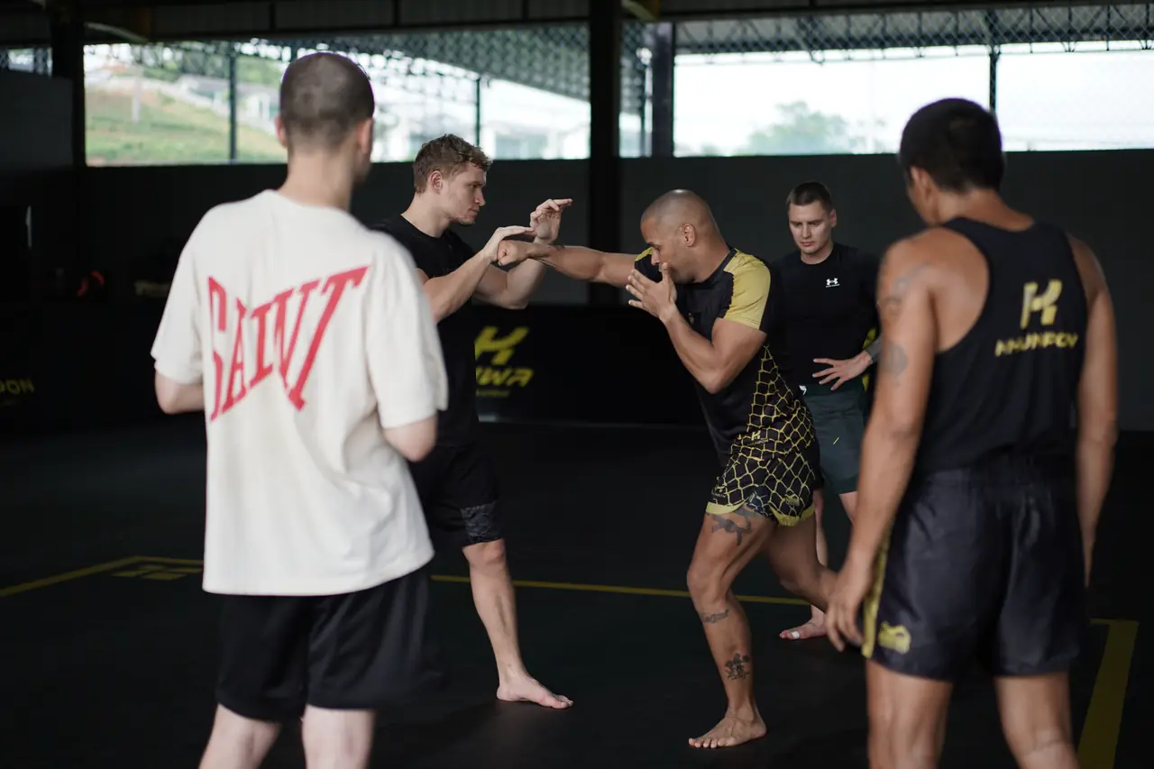 An MMA coach demonstrates a striking entry while fighters watch on the mats at Khunpon